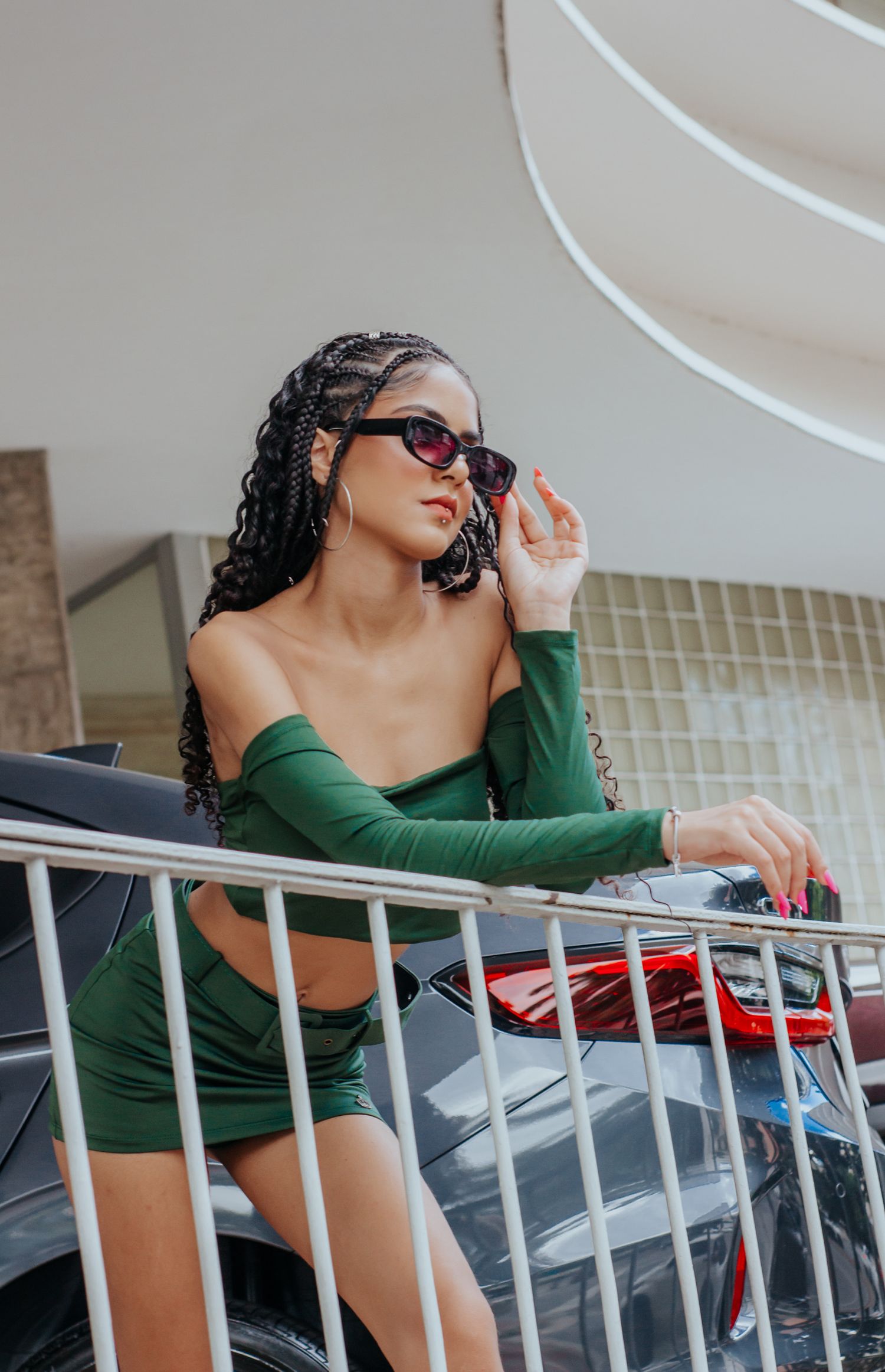 Fashionable woman in green outfit leaning on railing, showcasing urban style with sunglasses.